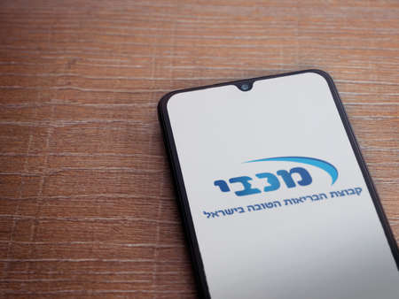 Lod, Israel - July 8, 2020: Maccbi Health Services app launch screen with logo in hebrew text on the display of a black mobile smartphone on wooden background. Top view flat lay with copy space.のeditorial素材
