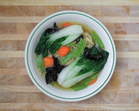 Vegetable noodle soup with bok choy, carrots, snow peas, and wood ear mushrooms in a white bowl on a wooden table.の写真素材