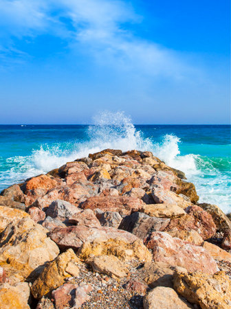 Seascape. Turquoise waves crashing on coastal rocks against blue sky with clouds.