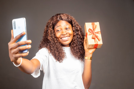 Portrait of a happy pretty African woman holding present box while standing and looking at phone camera isolated over gray studio backgroundの写真素材
