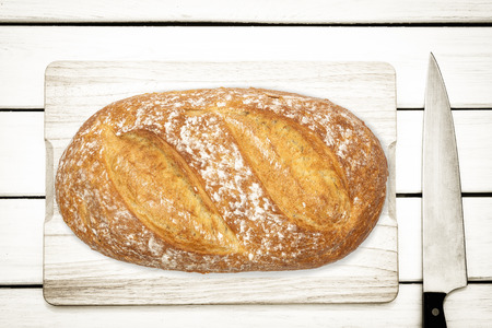 Rustic culinary concept - top view of a fresh homemade loaf bread and knife on a white wooden table.