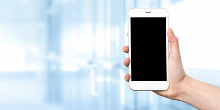 Woman hand holding a white smartphone with blank screen on a blurred background of a modern corporate interior with copy spaceの写真素材