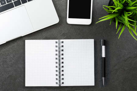 Top view of a dark office or home desk with laptop, smartphone, green plant and blank notebook with...