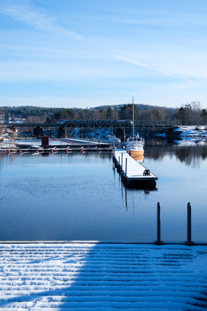 Snow blankets the banks of a serene waterfront in Leksand, Dalarna. Boats rest peacefully at the dock under a clear blue sky. A tranquil winter scene captures the essence of Swedish nature.の写真素材