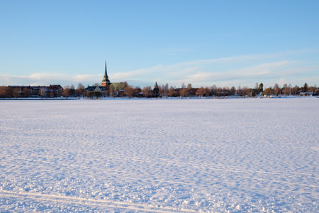 A serene winter landscape captured in Mora, Dalarna, Sweden. The scene showcases a vast snowy expanse with a church tower visible in the distance against a clear blue sky.の写真素材