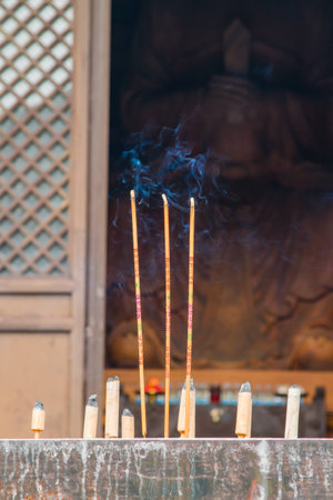 Burning incense at a stone alter in front of a temple housing a giant wooden sculpture of Buddha.の写真素材