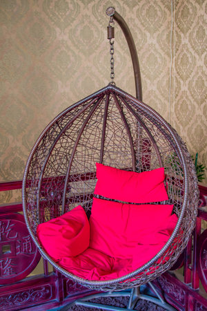A hanging wicker basket type chair with red cushions against an antique red wood fenceの写真素材