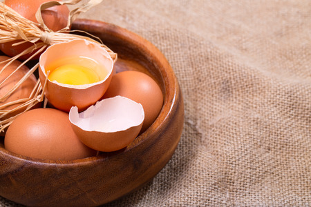 Wooden bowl with eggs, rural style. The bowl costs on a sacking, one egg is broken. Horizontal shot,...