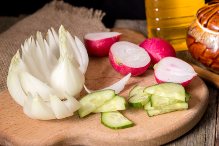 The cut vegetables on a wooden board. Onions, a garden radish, a cucumber, a bottle with oil. Close upの写真素材