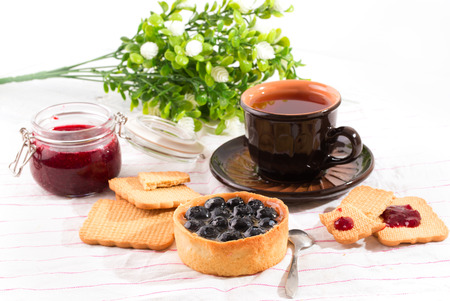 Breakfast with bilberry cake. Bilberry cake, cookies and a cup with tea on a light table. Flowers on a backgroundの写真素材