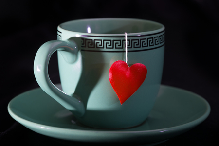 Cup with a shadow in the form of heart and a heart on a thread. A cup on a saucer, a red heart from a tea bag, a black backgroundの写真素材