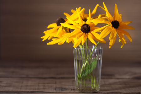 Yellow flowers of a gerbera in a glass. A bouquet from three flowers in a glass transparent thick glass tumbler on a wooden table. Close up, selective focus, free space leftの写真素材