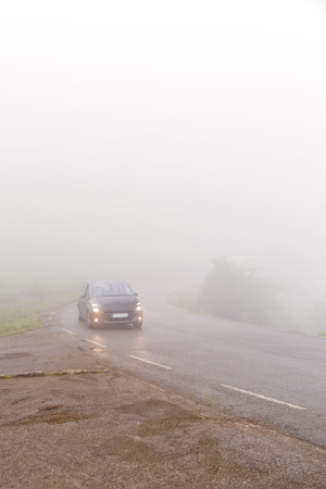 The car on the road in dense fog. Dense fog over the wet asphalted road in cloudy summer day, the car goes with the lit headlightsの写真素材