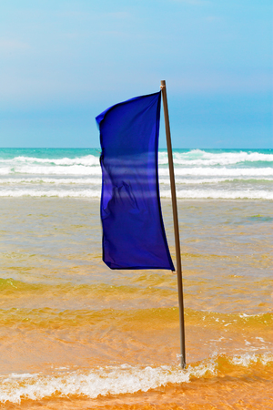 Blue flag on the beach. Shallow water, waves on the sea and the blue sky. Sunny summer day in Spainの写真素材