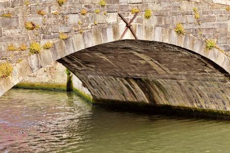 Arch of the ancient bridge from a stone. The bridge over the river with muddy green water, between stones of the bridge grows a grass.の写真素材