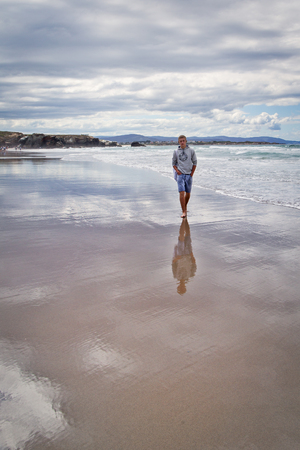 Playa de Las Catedrales,  Lugo, Galicia, Spain - July 12, 2016: The young man costs on the seashore. The barefoot young man in jeans shorts and a white linen bag on a shoulder costs on the sandy beach in a cloudy flying weather and looks afarのeditorial素材