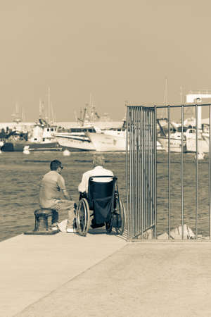 LLORET DE MAR,SPAIN - AUGUST 8, 2019: Two men sit on the edge of the pier on a summer day.のeditorial素材