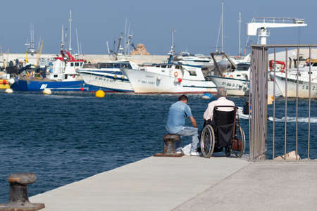 LLORET DE MAR,SPAIN - AUGUST 8, 2019: Two men sit on the edge of the pier on a summer day.のeditorial素材