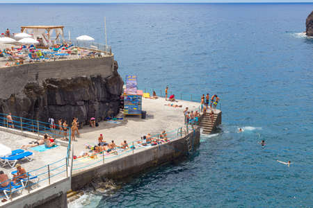 MADEIRA, PORTUGAL - JULY 28, 2018: Tourists sunbathe and bathe on the beach on a concrete pier in...