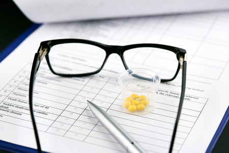 Glasses, yellow pills in graduated glass and pen on medical questionnaire form.の写真素材