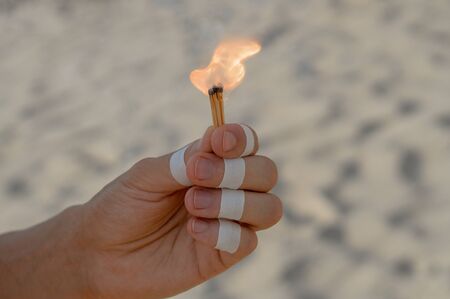 Hand with bandages holding a burning matchstick and sand background.