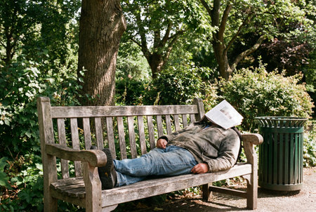 Tired man resting on park bench with newspaper covering face near trash canの素材