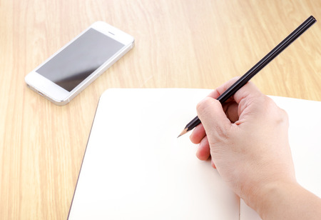 Hand holding black pencil and writing on blank open notebook with smartphone beside it on wooden table,Template mock up for adding your text.の写真素材