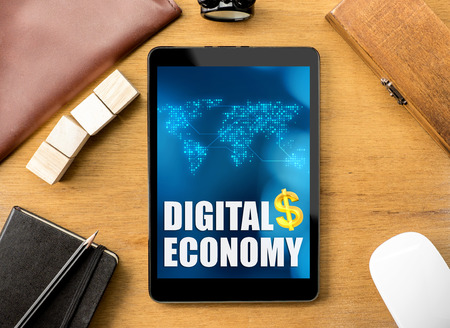 Digital Economy word on tablet with clock ,box,notebook,leather bag, pencil and mouse on wooden table, Digital marketing concept.の写真素材