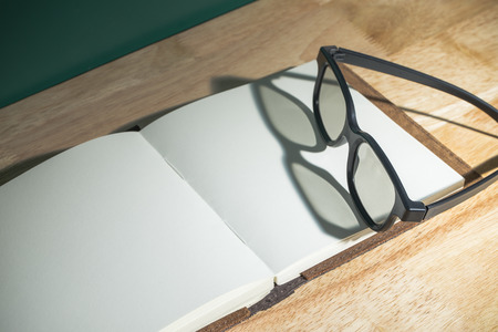Close up black eyeglasses on open blank book with shadow light from window on wooden table top and blackboard wall,Education concept,study resting.の写真素材