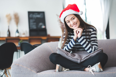 Asian teenage girl sitting cross-legged on sofa with Christmas hat and smile face and  resting her chin on her hands at home ,Celebrate New year and Christmas holiday concept.の写真素材