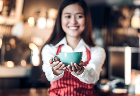 Close up on coffee up with blur asian woman barista holding mug with two hand with smiling face at...