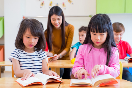 two Asian girl kid reading book in classroom and while teacher teach friends beside them,kindergarten education.の写真素材