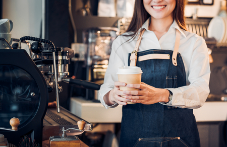 Close up on coffee take away cup with asian woman owner barista holding mug with two hand with smiling face at cafe bar background,Focus on coffee upの写真素材