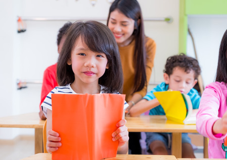 Asian girl kid reading book in classroom and while teacher teach friends beside them,kindergarten education preschool.の写真素材