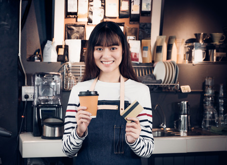 Woman barista holding coffee to go cup and credit card,asia female waitress using contactless payment in coffee shop business at counter bar in cafe,moder food owner business start upの写真素材