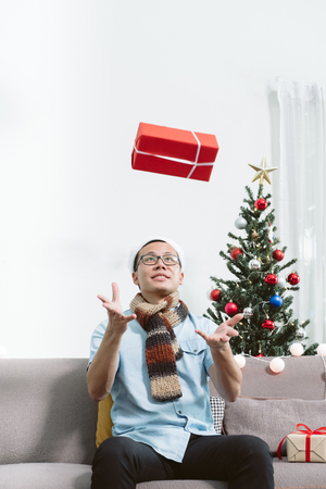 Asia man wear santa hat sit on sofa throw red gift box in air with happy emotion in christmas party at home,winter season holiday celebration.の写真素材