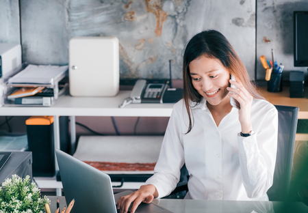 Asian businesswoman talking on mobile phone in front of laptop computer and looking screen at office,Office lifestyle conceptの写真素材
