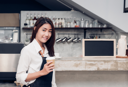 Asian female barista wear jean apron holding take away cup at machine counter bar with smile face,cafe service concept.making coffeeの写真素材