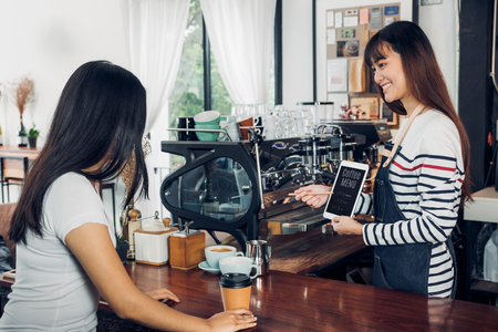 Asian woman barista wear jean apron suggestion coffee menu to customer at bar counter with smile emotion,Cafe restaurant service concept,Owner small business concept.の写真素材