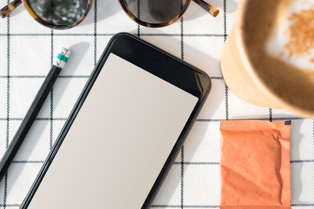 Top view of mobile phone with blank screen with coffee cup,sunglasses,pencil on white table...