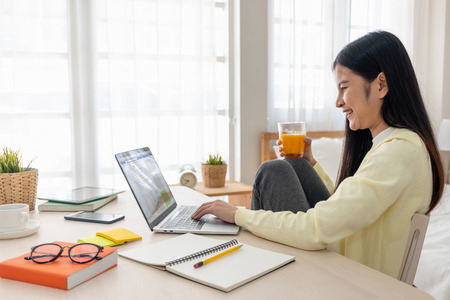 Asian female sit with knees up use social media with laptop on table and drink orange juice in bedroom at home.Work at home concept.work from home.relax lifestyleの写真素材