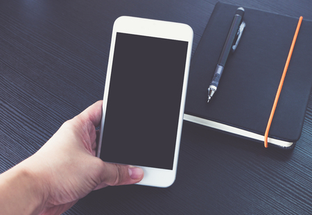 Hand holding Blank screen of mobile phone over black notebook on black wooden office desk, Mock up for display of design or content,Clipping path smart phone screenの写真素材