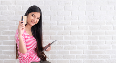 Asian woman in casual cloth holding credit card and using to online shopping with tablet at white brick wall.Digital technology lifestyle.Banner with copy space for display text or designの写真素材