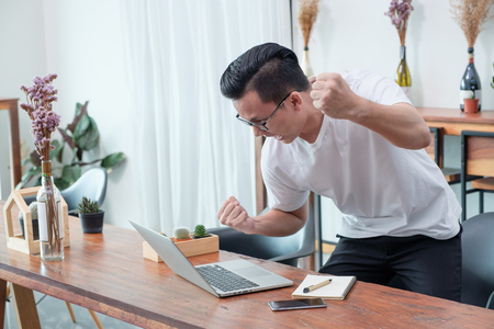 asian casual man standing and arm up celebrate good new from mobile at laptop in coffee shop,success business concept,work outside office,work at homeの写真素材
