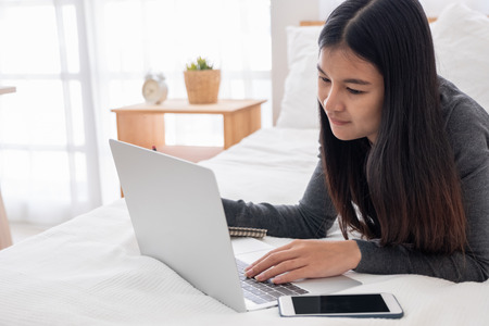 Asian woman laying on bed doing paperwork on laptop in bedroom.work at home.e-larning concept.study onlineの写真素材