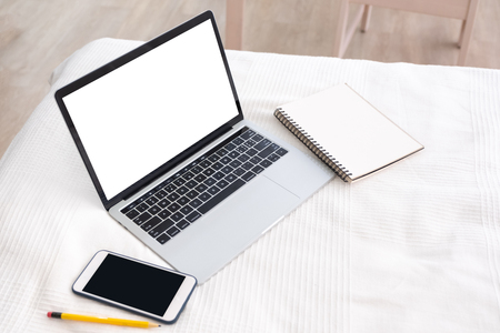 Mock up white screen of laptop with mobile phone and blank notebook on bed blanket in bedroom.working at  home concept.copy space for adding design or text with clipping pathの写真素材