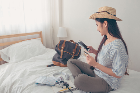 Young asian woman traveller planning vacation holiday expedition and booking ticket and hotel online with mobile phone app on bed.preparing to travel.digital lifestyleの写真素材