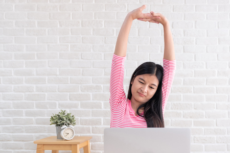 Asian woman stretching arm up when working on laptop at white brick wall in home.Work at home concept.relax after hardworkの写真素材