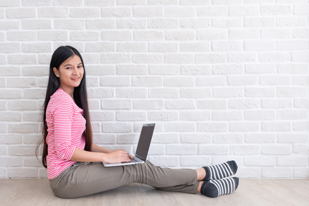 Asian woman in casual pink cloth use laptop and sitting on floor at white brick wall in house.mock up screen and copy space for adding text or designの写真素材
