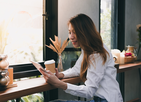 Asian Businesswoman holding coffee cup and reading report on tablet computer on wood stool table at coffee shop near window,woking outside office lifestyleの写真素材